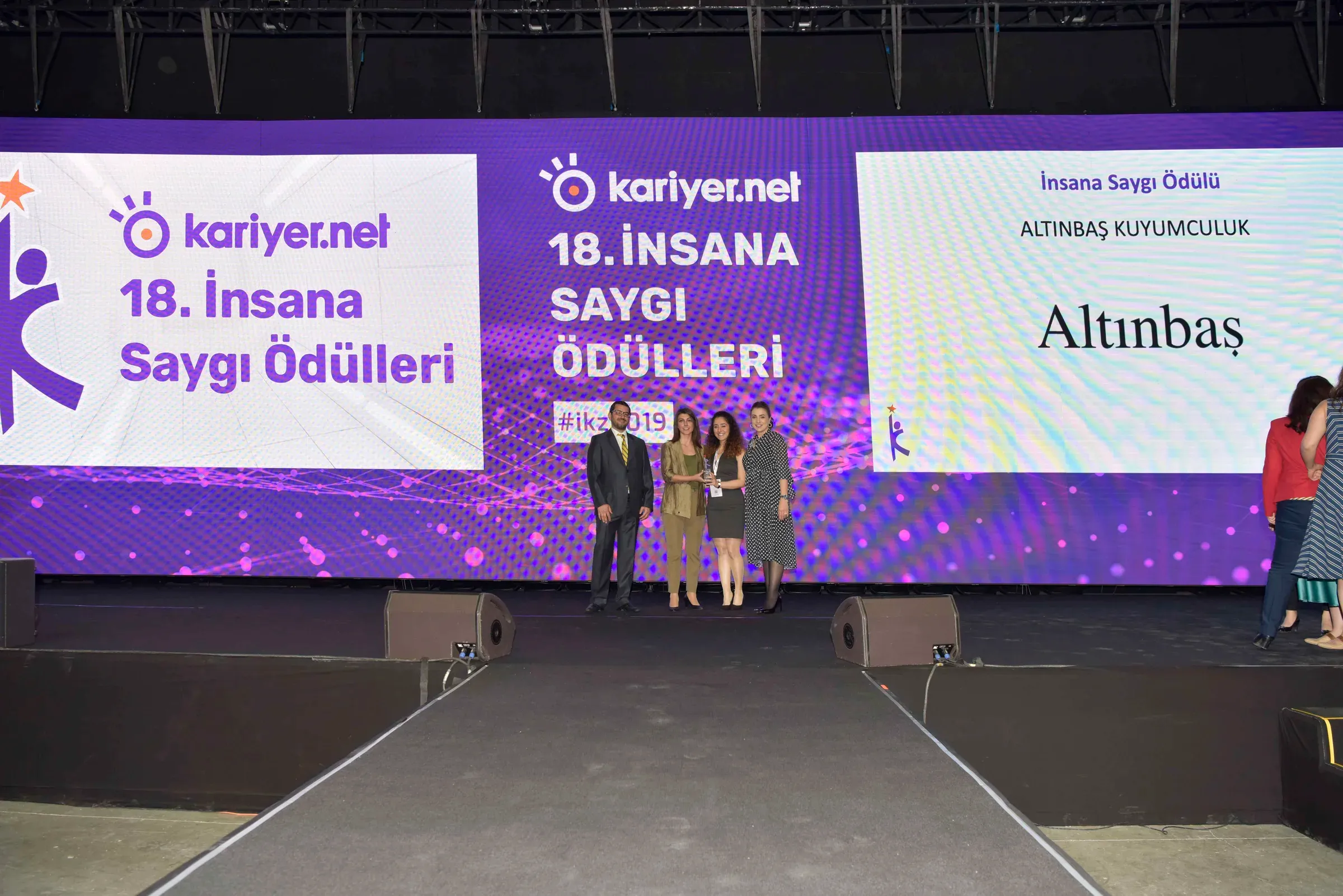 Ödül sahnesinde büyük ekran önünde duran katılımcılar.